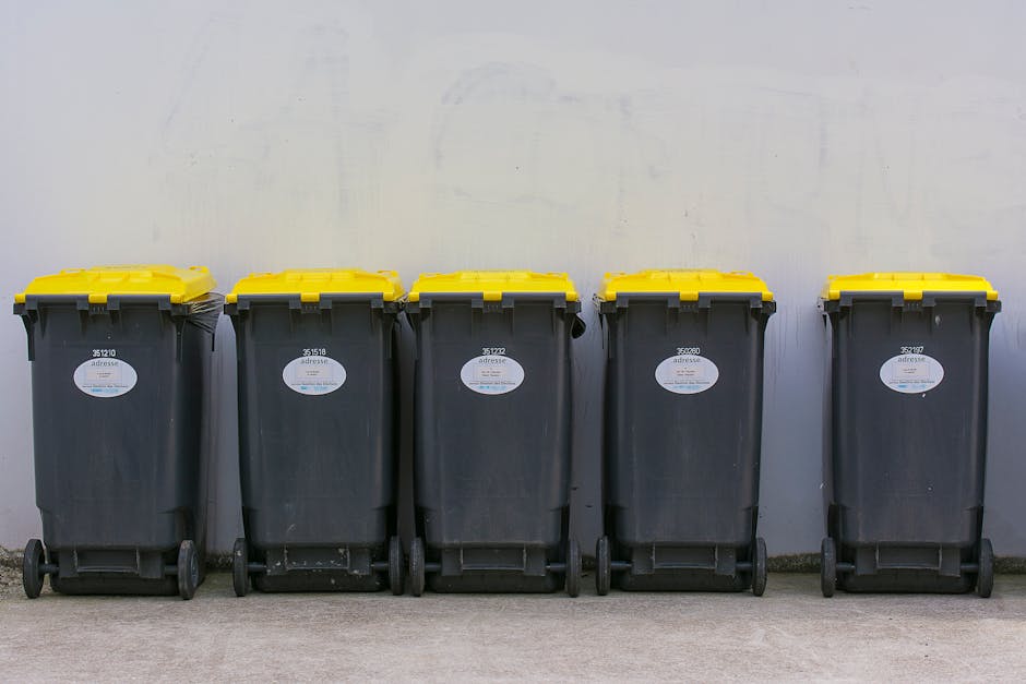 A row of five large wheeled rubbish bins made of black plastic with yellow lids positioned against a plain, off-white wall. Each bin has a white oval label with black text and a barcode, and they are 