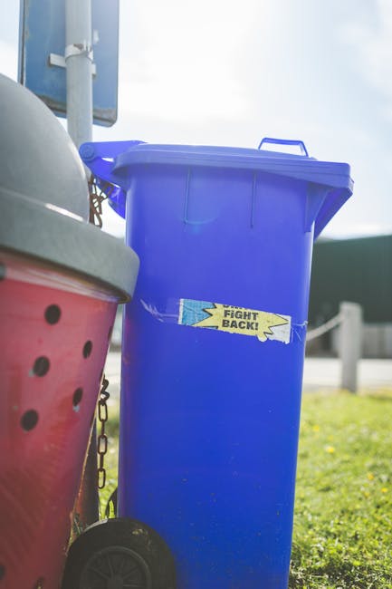A row of five large wheeled rubbish bins made of black plastic with yellow lids positioned against a plain, off-white wall. Each bin has a white oval label with black text and a barcode, and they are 