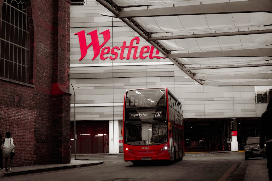 A red double-decker bus is seen entering or exiting a parking area in front of a large modern shopping centre with a prominent white facade and the bold red signage displaying 'Westfield'. The bus dis